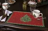 A Christmas-themed placemat with a decorated Christmas tree is set on a dark wooden table, with a mug, napkin, and cutlery. Produktbild-Vorschau