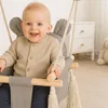 A baby is sitting in a baby swing in a room with a dresser and a stuffed bunny in the background. Náhled obrázku produktu