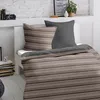 A neatly made bed with striped bedding and a dark gray pillow, set against a white brick wall. Produktbild-Vorschau