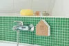 A bathroom with green tiled walls, a chrome faucet, and a small house-shaped speaker with a wood-grain front. Produktbild-Vorschau