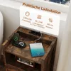A close-up shot of a rustic brown nightstand with electronic devices charging on top and a decorative item below. Produktbild-Vorschau