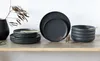 A set of dark gray plates and bowls are neatly stacked on a wooden shelf, with a decorative dried plant branch and a black vase in the background. Podgląd zdjęcia produktu