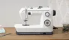 A white Husqvarna Viking sewing machine is displayed on a wooden table with some sewing supplies in the background. Produktbild-Vorschau