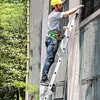 A person wearing a yellow hard hat and a safety harness is climbing a silver multi-purpose ladder against a building. Náhľad obrázku produktu