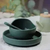 A green silicone baby bowl and plate set with a spoon, placed on a textured surface with blurred greenery in the background and a light-colored teapot to the right. Náhľad obrázku produktu