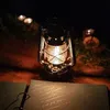 A petroleum lantern is lit and placed on a wooden surface next to an open book with handwritten text. Náhľad obrázku produktu