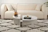 A modern living room scene featuring a cream-colored sofa, a white coffee table with a book and candle, and a textured rug. Produktbild-Vorschau