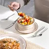 A person is eating a bowl of fruit and oatmeal with a spoon, with a plate of pasta and cutlery on the table. Produktbild-Vorschau