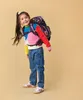 A young girl wearing a colorful sweater and jeans, with a backpack on, posing against a plain background. Náhľad obrázku produktu