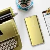 A gold smartphone with a clear view flip case is placed on a white surface next to a vintage typewriter and a small potted plant. Produktbild-Vorschau