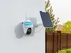 A security camera and solar panel mounted on a white wall, with a fence and trees in the background. Produktbild-Vorschau