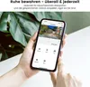 A person is holding a smartphone displaying a security camera app, with a large green leaf in the background. Náhled obrázku produktu