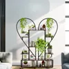 A heart-shaped plant stand filled with various potted plants and books, placed in a well-lit room with a sofa visible. Produktbild-Vorschau