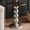 A round wooden-topped side table with a stacked silver ball pedestal base, placed on a wooden floor next to a sofa and a woven basket. Voir les images du produit