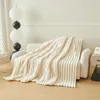 A cream-colored plush blanket is draped over a modern sofa, with a floor lamp in the background. Produktbild-Vorschau