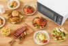 A variety of food dishes are arranged on a wooden table next to a white microwave. Podgląd zdjęcia produktu