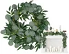 A wreath and garland made of artificial eucalyptus leaves are shown, with the garland draped over a white brick fireplace decorated with candles. Anteprima dell’immagine prodotto