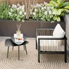 A modern outdoor seating area with a black metal couch and a small black side table on a circular rug, surrounded by lush greenery and white flowers. Anteprima dell’immagine prodotto