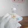 A baby bottle with milk and a pacifier are placed on a white cloth next to a wooden surface. Produktbild-Vorschau