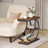 A side table with shelves next to a cream-colored armchair, with a vase of flowers and a cup on top. Produktbild-Vorschau