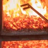 A close-up view of burning wood chips and embers in a furnace or fireplace, with a metal tool partially visible. Produktbild-Vorschau