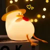 A cute, glowing duck-shaped night light wearing a hat and holding a red heart, with yellow hearts and bokeh lights in the background. Produktbild-Vorschau