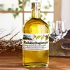 A bottle of Maikäferflugbenzin liqueur sits on a wooden surface, with glasses in the background and a window with greenery behind it. Produktbild-Vorschau
