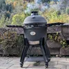 A black kamado grill is shown outdoors on a patio with a stone wall and foliage in the background. Produktbild-Vorschau