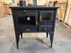 A black wood-burning stove with an oven and metal plate is displayed in a warehouse or storage area. Anteprima dell’immagine prodotto
