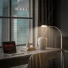 A desk with a laptop, books, a vase with branches, and a floor lamp, set against a window with a city view. Náhľad obrázku produktu
