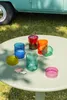 A collection of colorful glass jars and lids are arranged on a light green table outdoors, with grass and a teal vehicle in the background. Produktbild-Vorschau