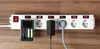 A white power strip with multiple outlets and individual switches is mounted on a wooden surface. Several devices are plugged into the power strip, including a battery charger with batteries, and a black power adapter. Produktbild-Vorschau