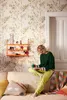 A woman sits on a sofa, reading a book and holding a mug, in a room with floral wallpaper. Produktbild-Vorschau