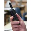 A hand holding a black walkie-talkie in a classroom or office setting. Produktbild-Vorschau