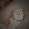 A person holding a small, white, oval-shaped device with a glowing orange light in the center, while lying in bed. Podgląd zdjęcia produktu