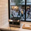 A waffle maker is shown on a counter in front of a window looking out onto a street with pedestrians. Podgląd zdjęcia produktu