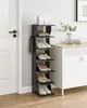 A black multi-tiered shoe rack filled with various shoes and a white handbag on top, situated next to a white door. A cabinet and a plant are visible in the background. Náhled obrázku produktu