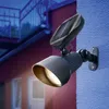 A solar-powered spotlight mounted on a blue wall, with a red building in the background. Produktbild-Vorschau
