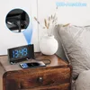 A digital alarm clock with a projected time is shown on a nightstand next to a bed, with a smartphone charging. Produktbild-Vorschau