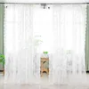 A close-up view of white lace curtains with a floral embroidery pattern, partially obscuring a window and a wooden table with plants. Produktbild-Vorschau