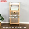 A bamboo shelving unit with a laundry basket integrated into the bottom cabinet, shown in a room setting with a plant. Náhled obrázku produktu