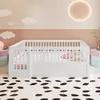 A white wooden crib with a door and railing, set up in a child's room with cloud decorations on the wall and a patterned rug on the floor. Produktbild-Vorschau