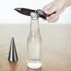 A hand is using a black bottle opener to open a clear glass bottle on a wooden table. A cone-shaped object is to the left of the bottle. Podgląd zdjęcia produktu