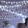 A string of fairy lights with photos clipped to them, hanging on a wall above a table with a plant and a chair. Produktbild-Vorschau