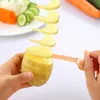A potato is being spiralized with a tool, with sliced carrots and cucumbers in the background. Náhled obrázku produktu