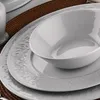 A close-up view of white porcelain dinnerware, including a large oval serving bowl and a plate with embossed decorative patterns, set on a brown woven placemat. Produktbild-Vorschau