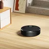 A black robot vacuum cleaner is on a light wood floor next to a white cabinet and a framed picture. Stairs are visible in the background. Produktbild-Vorschau