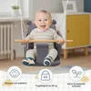 A smiling baby sits in a gray swing with a wooden bar, indoors. Produktbild-Vorschau