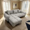 A light gray sectional sofa is placed in a well-lit living room with large windows and sheer curtains. Produktbild-Vorschau
