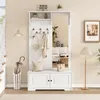 A white entryway hall tree with a mirror, coat hooks, a bench with cabinet doors, and a shelf above. It is set in a home entryway with a staircase, a floor lamp, and a framed picture on the wall. Produktbild-Vorschau